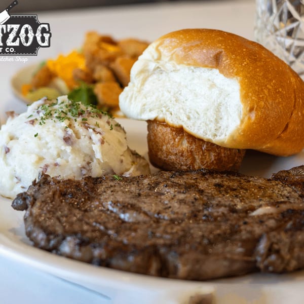 A grilled steak, mashed potatoes, and a dinner roll on a white plate with a glass of water.