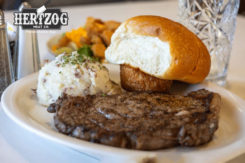A grilled steak, mashed potatoes, and a dinner roll on a white plate with a glass of water.